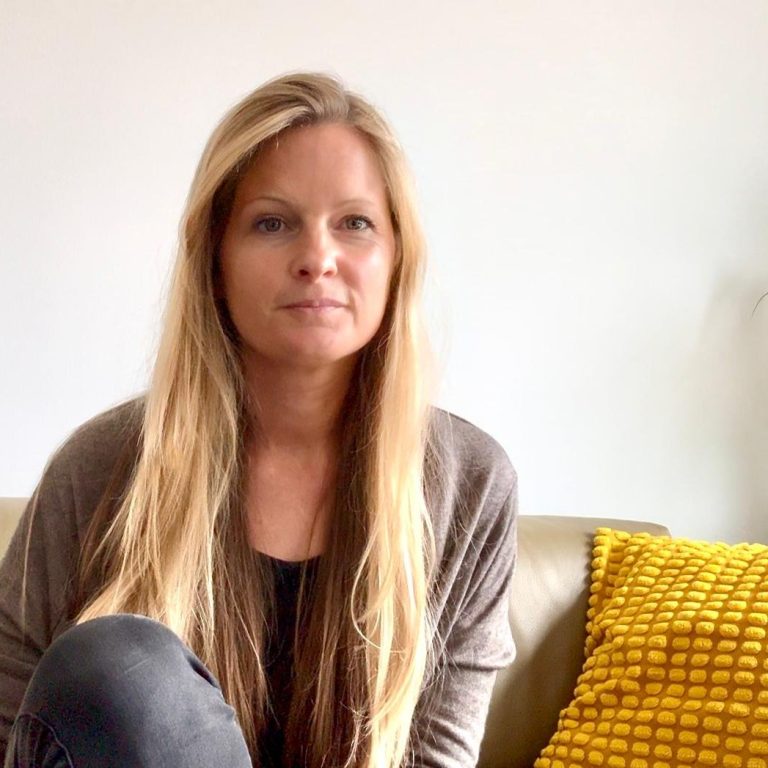 Inga Mehlhardt mit langen, blonden Haaren sitzt auf einer Couch neben einem gelben Kissen.