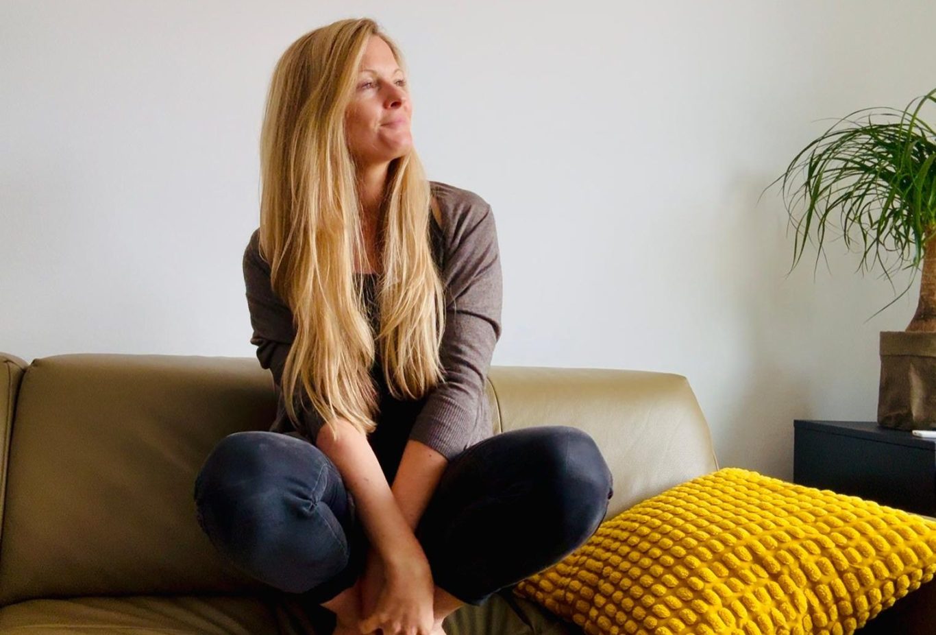 Feinfühlige junge Frau mit langen Haaren sitzt auf einer Couch und blickt ruhig zur Seite – Symbol für leise Stärke und inneres Wachstum.